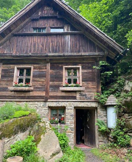 Ein altes Holzhaus, datiert 2003, mit Blumenkästen an den Fenstern und einer Person vor der Tür.