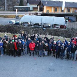 Eine Gruppe von Menschen in Winterkleidung posiert für ein Foto draußen vor einem Parkplatz mit Autos. Dahinter stehen mehrere Gebäude mit geparkten Autos vor ihnen.