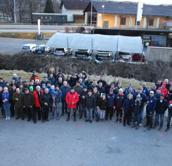 Eine Gruppe von Menschen in Winterkleidung posiert für ein Foto draußen vor einem Parkplatz mit Autos. Dahinter stehen mehrere Gebäude mit geparkten Autos vor ihnen.