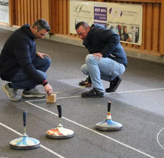 Zwei Männer spielen eine Partie Curling auf einem Betonboden. Ein Mann kniet und der andere steht, beide schauen auf ein Ziel hinunter.