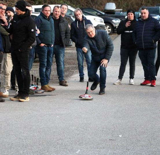 Eine Gruppe von Männern steht um einen Mann herum, der auf einer Straße einen Curlingstein wirft, während andere zuschauen.