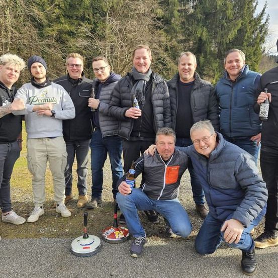 Eine Gruppe von Männern in lässiger Kleidung posiert für ein Foto im Freien. Sie lächeln und halten Bierflaschen in den Händen. Ein Mann kniet und hält eine Bierflasche. Sie stehen vor Bäumen und einer Betonfläche.