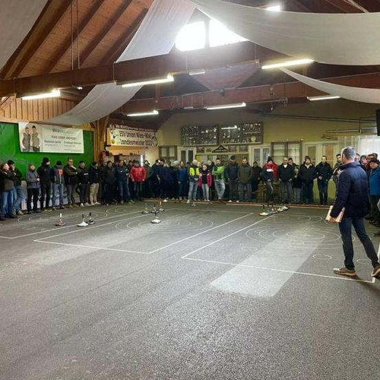 Eine Menschenmenge versammelt sich in einem Saal mit einem Basketballplatz-Setup. Eine Person geht vorwärts und hält ein Buch. Banner hängen an den Wänden, und Lampen sind an der Decke montiert.