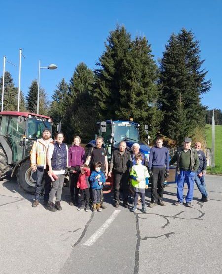 Eine Gruppe von Menschen, Erwachsenen und Kindern, steht vor zwei Traktoren, mit Bäumen im Hintergrund, an einem sonnigen Tag.