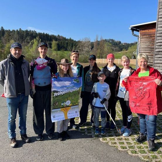 Eine Gruppe von Menschen steht an einem sonnigen Tag vor einem Gebäude. Sie halten ein Plakat und einen roten Stoff und scheinen für ein Foto zu posieren. Bäume und Berge sind im Hintergrund.