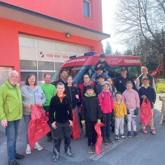Eine Gruppe von Menschen, darunter Kinder, steht vor einer Feuerwache. Einige halten rote Tüten. Ein Feuerwehrauto ist in der Nähe geparkt.