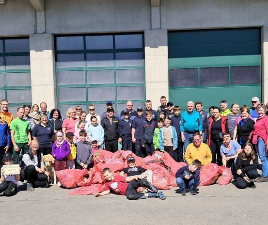 Eine Gruppe von Menschen, darunter Erwachsene und Kinder, posiert für ein Foto vor einem Gebäude mit grünen Garagentoren und hält rote Taschen, einige mit Hunden.