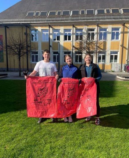 Drei Personen, wahrscheinlich Studenten, stehen auf einem grünen Rasen und halten rote Tüten mit gedruckten Botschaften. Sie stehen vor einem gelben Gebäude mit Solarmodulen auf dem Dach.