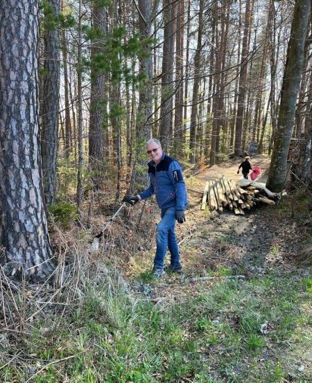 Ein Mann in einer blauen Jacke und Jeans trimmt Äste mit einer Kettensäge in einem Kiefernwald. Im Hintergrund geht eine Person mit einem Kind auf einem Pfad.
