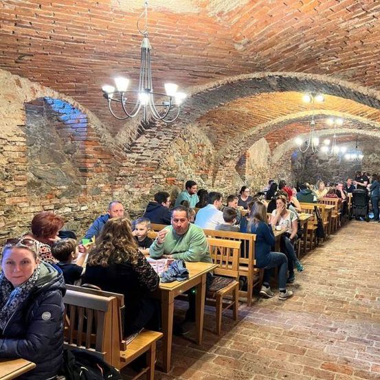 Eine Gruppe von Menschen speist in einem Kellerrestaurant mit gemauerten Wänden, gewölbten Decken und Kronleuchtern.