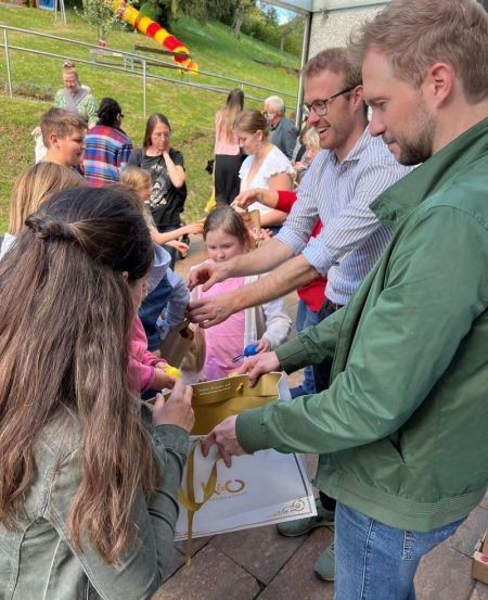 Eine Gruppe von Erwachsenen und Kindern versammelt sich um einen Tisch und erhält Gegenstände aus einer Kiste, mit einem Grashügel und einem Spielplatz im Hintergrund.