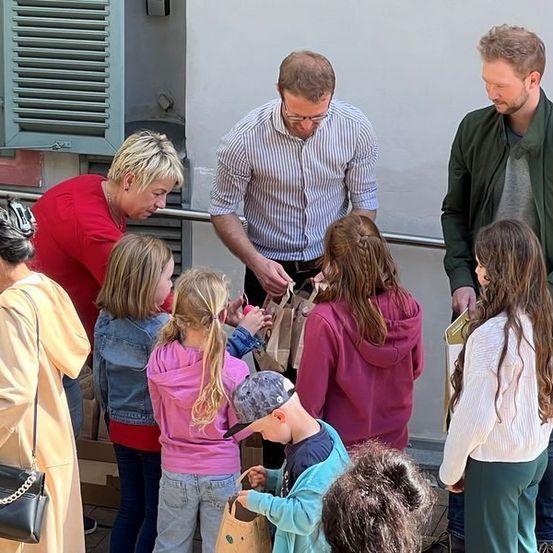 Mehrere Erwachsene und Kinder versammeln sich draußen, möglicherweise in einer Schulumgebung. Zwei Männer mit Brille und Jacken verteilen Gegenstände in Papiertüten an die Kinder.