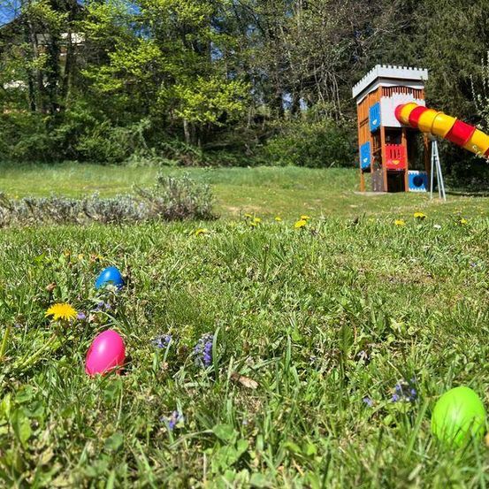 Eine Ostereiersuche in einem Park mit einer Rutsche und einem Spielhaus. Drei Eier sind im Gras verstreut.