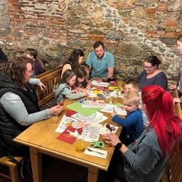 Eine Gruppe von Erwachsenen und Kindern sitzt um einen Holztisch in einem Raum mit Steinwänden und bastelt.