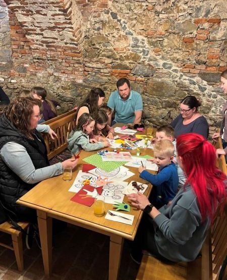 Eine Gruppe von Erwachsenen und Kindern sitzt um einen Holztisch in einem Raum mit Steinwänden und bastelt.