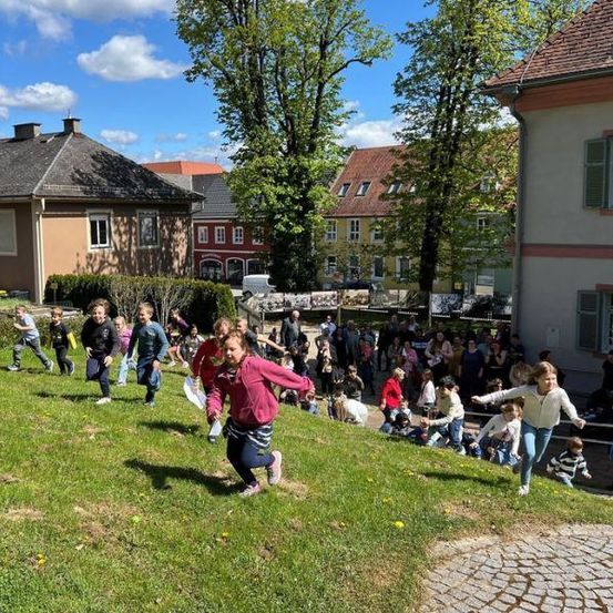 Kinder laufen auf einem grasigen Hügel, möglicherweise bei einer Ostereiersuche, mit Gebäuden und Menschen im Hintergrund.