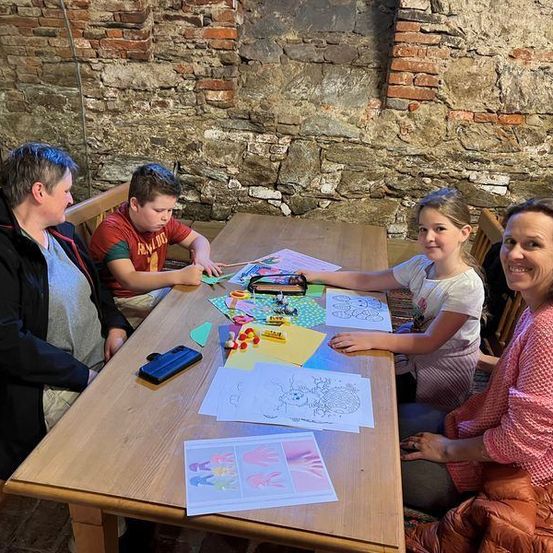 Vier Personen sitzen um einen Holztisch herum und sind in einer Malaktivität beschäftigt. Zwei Frauen und zwei Kinder, ein Junge und ein Mädchen, sind anwesend. Sie alle lächeln und scheinen sich zu amüsieren. Der Tisch ist mit verschiedenen Malmaterialien bedeckt, und das Mädchen hält einen Marker. Hinter ihnen ist eine Steinmauer.