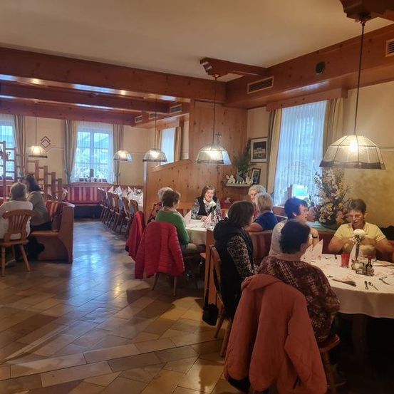 Ein Innenrestaurant mit Holzwänden und Decken, in dem eine Gruppe von Menschen an Tischen mit weißen Tischdecken sitzt, sich unterhält und isst.