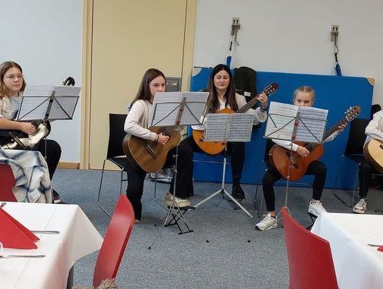 Eine Gruppe junger Frauen sitzt auf Stühlen mit Notenständern und spielt Gitarre. Sie befinden sich in einem Raum mit Tischen, die mit weißen Tischdecken und roten Servietten bedeckt sind. Ein Glas Orangensaft steht auf einem Tisch.