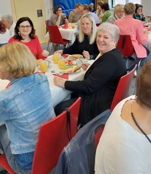 Mehrere Frauen sitzen an einem Esstisch, einige lächeln und interagieren. Sie haben Teller mit Essen, Gläser Orangensaft und Servietten. Der Raum hat eine warme, gelbe Beleuchtung.