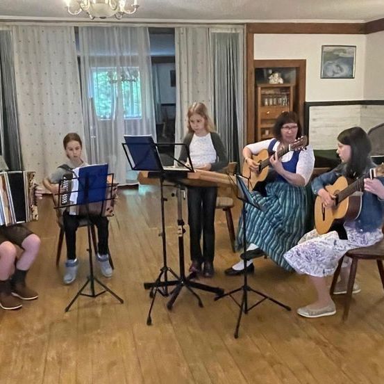 Fünf Kinder spielen Instrumente in einem Raum. Ein Junge spielt Akkordeon, während die Mädchen Gitarre spielen. Sie haben Notenständer vor sich. Dahinter steht eine Frau, die ebenfalls Gitarre spielt.