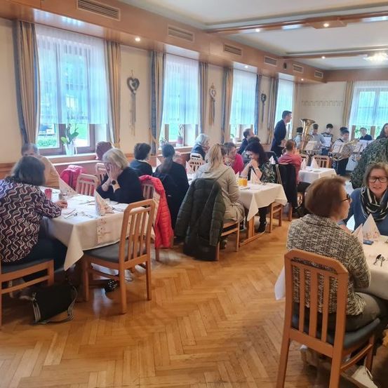 Eine Gruppe Erwachsener sitzt an Tischen in einem Essbereich mit Holzboden, weißen Tischdecken und großen Fenstern. Einige Leute stehen im Hintergrund und Musikinstrumente sind sichtbar.