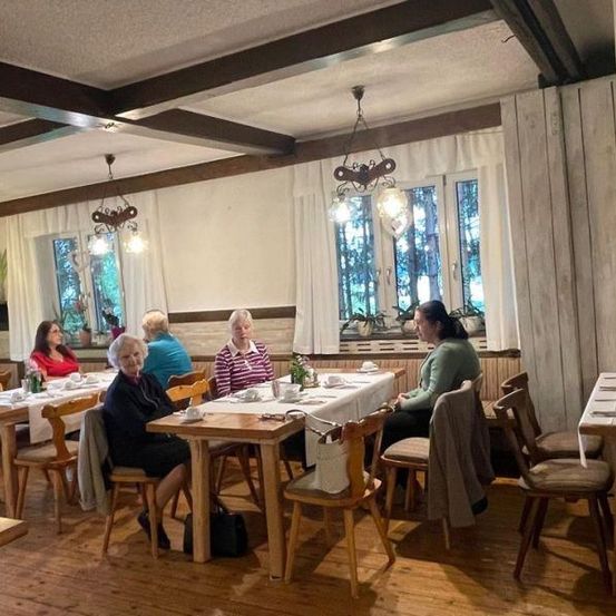 Ein Esszimmer mit mehreren älteren Frauen, die an Tischen sitzen. Der Raum hat Holzböden, weiße Vorhänge und hängende Lampen. Jeder Tisch ist mit Tassen, Untertassen und Besteck gedeckt.