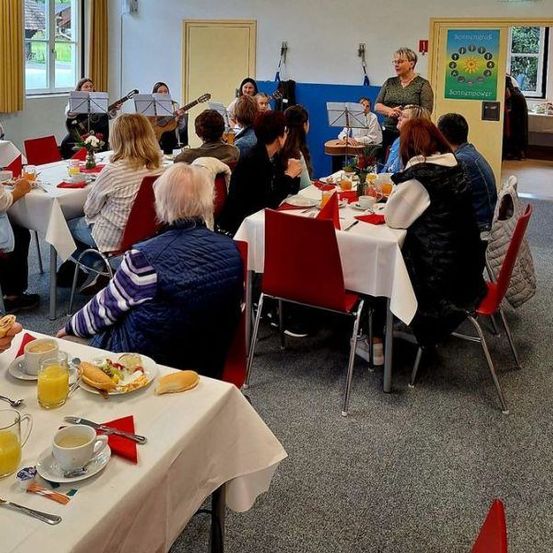 Eine Gruppe von Menschen ist in einem Essbereich versammelt. Einige sitzen, andere stehen in der Nähe einer gelben Wand, mit einer Frau vor einem Podium. Musikinstrumente und Frühstücksgegenstände sind vorhanden.