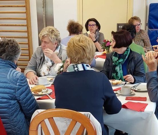Eine Gruppe von Frauen sitzt um einen Tisch und genießt eine Mahlzeit. Einige tragen Brillen und einige halten Tassen. Ein Blumenstrauß steht auf dem Tisch und Servietten liegen vor ihnen.