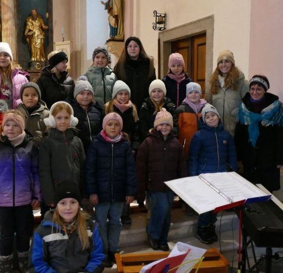 Eine Gruppe von Kindern und Erwachsenen steht in einer Kirche mit einem Klavier und Notenblättern vor ihnen. Sie sind alle warm für kaltes Wetter gekleidet.