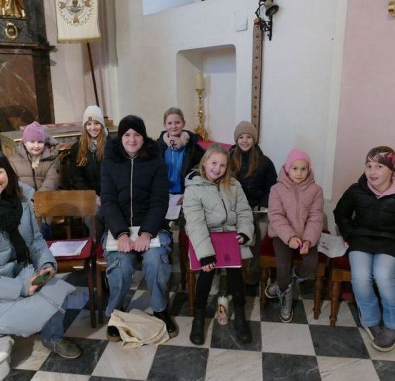 Eine Gruppe von Menschen sitzt in einer Kirche, einige in Winterkleidung. Sie lächeln und posieren für ein Foto. Eine Frau hält ein Buch.
