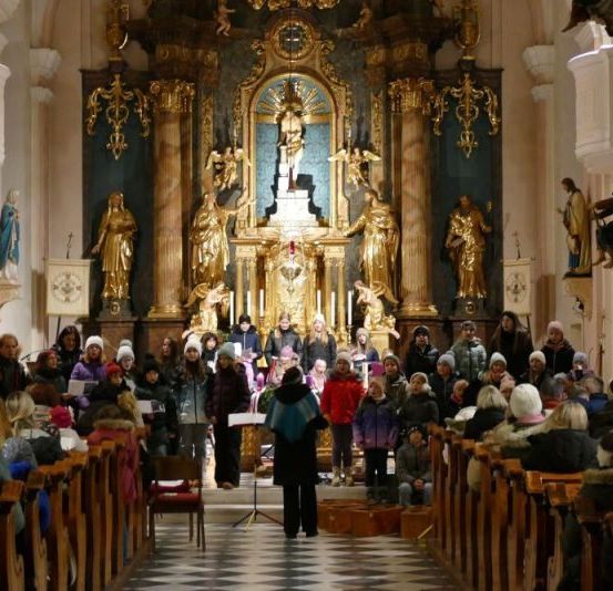 Ein Chor steht vor einem Kirchenaltar, mit Menschen, die auf den Kirchenbänken sitzen. Der Altar hat goldene Statuen, eine zentrale Jesus-Statue und ein goldenes Kreuz. Der Raum ist gut beleuchtet, und alle sind warm gekleidet.
