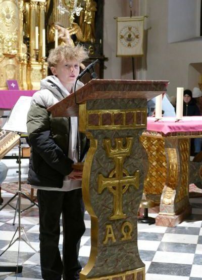 Ein junger Junge steht an einem Rednerpult mit Mikrofon und hält eine Rede in einer Kirche. Ein Kreuz befindet sich auf dem Podest, und hinter ihm stehen zwei Kerzen auf einem Tisch.