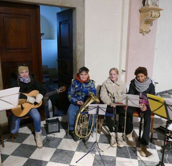 Eine Gruppe von Kindern spielt verschiedene Musikinstrumente in einer Kirche, darunter Gitarre, Tuba, Klarinette und Akkordeon.