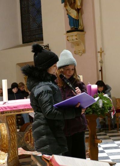 Zwei Mädchen stehen vor einem Kirchenaltar, halten ein Mikrofon und eine lila Mappe. Hinter ihnen ist eine Statue an der Wand befestigt, und ein Tisch mit Kerzen ist aufgestellt. Eine Person sitzt rechts auf einer Bank.