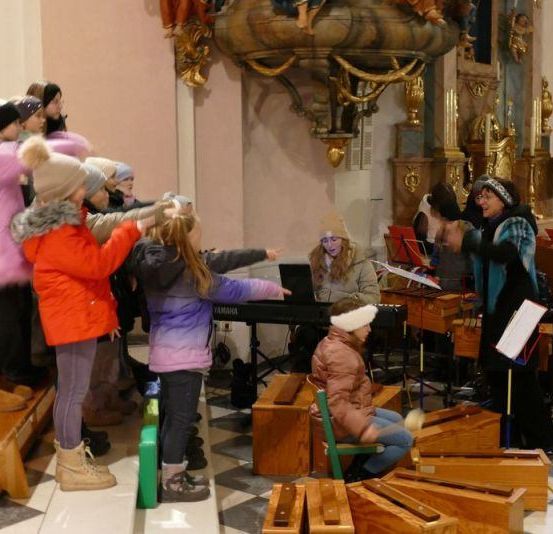 Eine Gruppe von Kindern und Erwachsenen steht und sitzt in einer Kirche. Sie sind warm gekleidet. Einige spielen Instrumente.