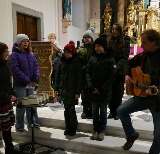 Eine Gruppe von Kindern und Erwachsenen steht auf den Stufen einer Kirche, ein Mann spielt Gitarre und andere sehen zu.