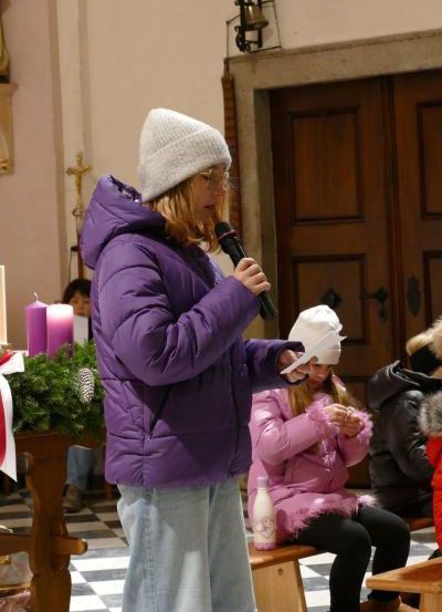 Eine Frau in einer lila Jacke steht in einer Kirche am Mikrofon und spricht zu einem sitzenden Kind in einer rosa Jacke und anderen. Hinter ihnen stehen brennende Kerzen, ein Kranz und eine Holztür.
