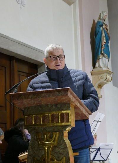 Ein älterer Mann steht in einer Kirche an einem Podest, trägt eine blaue Jacke und eine Brille. Eine Statue der Jungfrau Maria steht hinter ihm, und ein Mikrofon befindet sich auf dem Podest. Eine Frau im Hintergrund hält eine Gitarre.