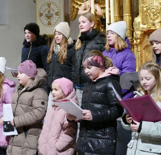 Eine Gruppe junger Mädchen, die Wintermäntel und Mützen tragen, singen in einer Kirche. Sie halten Papiere in der Hand und tragen verschiedenfarbige Mützen. Hinter ihnen steht ein goldener Altar.