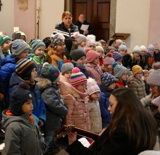 Eine Gruppe von Kindern steht und schaut nach vorne, wahrscheinlich in einer Kirche. Eine Person hält eine Gitarre und scheint zu spielen. Eine Person steht neben ihnen und hält ein Papier in der Hand. Einige Leute stehen hinter ihnen und einige sitzen. Ein Kreuz ist an der Wand hinter ihnen angebracht.