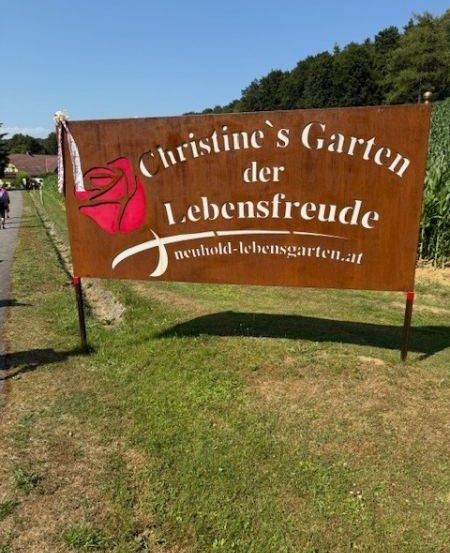 Ein braunes Schild von Christines Garten des Lebens mit einer roten Rose und einer Website-Adresse steht auf einem Grasbereich. Eine Person geht auf einem Weg neben einem Maisfeld.