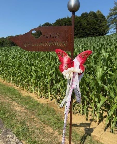 Ein Metallpfahl mit einem Schmetterling steht in einem Maisfeld. Ein Schild steht 'LICH WILLKOMMEN'. Bäume und Büsche sind im Hintergrund.