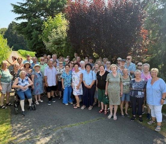 Eine große Gruppe von Menschen, einschließlich Senioren, versammelt sich für ein Foto in einem Garten mit Bäumen und Büschen. Einige tragen Hüte, Brillen und Turnschuhe.