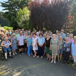 Eine große Gruppe von Menschen, einschließlich Senioren, versammelt sich für ein Foto in einem Garten mit Bäumen und Büschen. Einige tragen Hüte, Brillen und Turnschuhe.