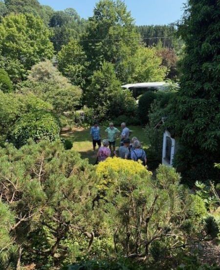 Eine Gruppe von Menschen steht in einem Garten mit verschiedenen Pflanzen und Bäumen. Ein kleines Gebäude ist rechts sichtbar.