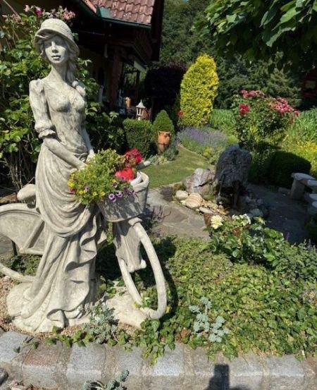 Eine Statue einer Frau mit Blumen steht auf einem Fahrrad in einem Garten mit verschiedenen Pflanzen und Bäumen.