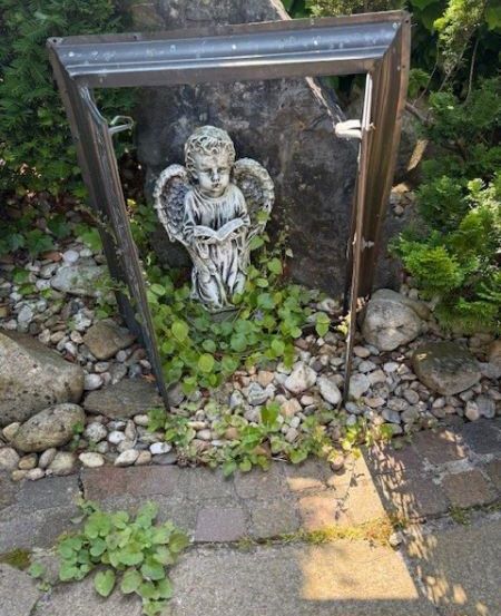 Eine Gartenszene mit einer kleinen Engelsstatue, die ein Buch in der Hand hält, unter einem Fensterrahmen in der Nähe eines großen Felsens platziert.