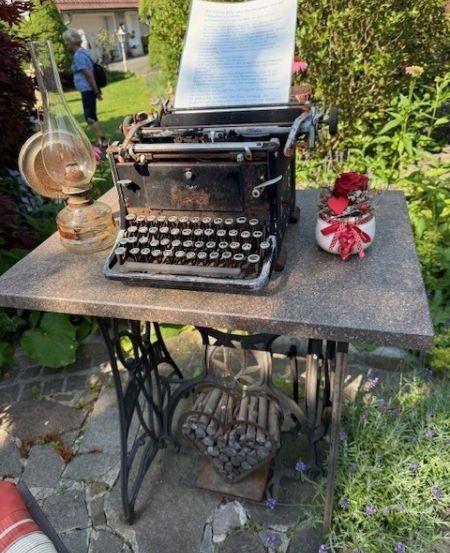 Eine alte Schreibmaschine steht auf einem Tisch in einem Garten mit einer Lampe und einem Blumentopf. Eine Person geht im Hintergrund.