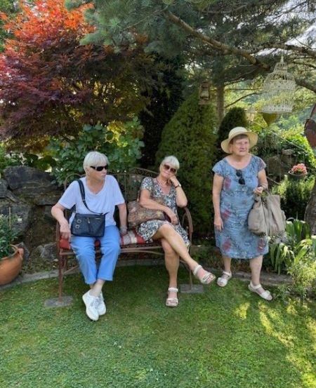 Drei Frauen sind in einem Garten. Eine Frau sitzt auf einer Bank und die anderen beiden stehen. Sie tragen lässige Sommerkleidung. Hinter ihnen befinden sich verschiedene Pflanzen und Bäume.